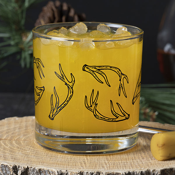 Whiskey glass printed with black antlers, filled with ice and an orange colored drink..