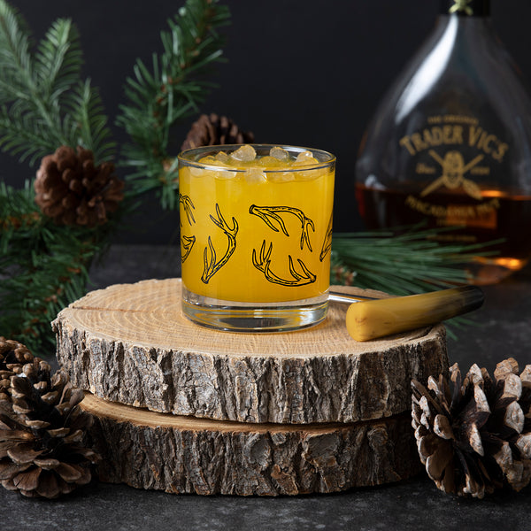 Whiskey glass printed with black antlers, filled with ice and an orange colored drink.