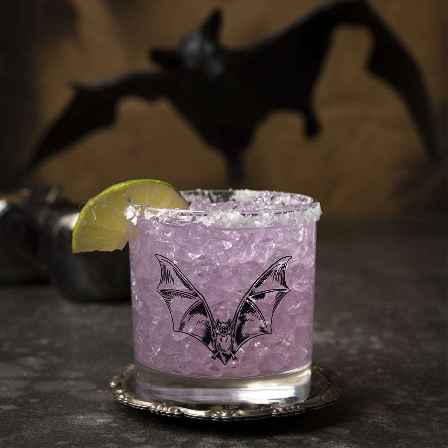 Bat whiskey glass with purple drink, ice, and lime garnish. Spooky bat in the background.