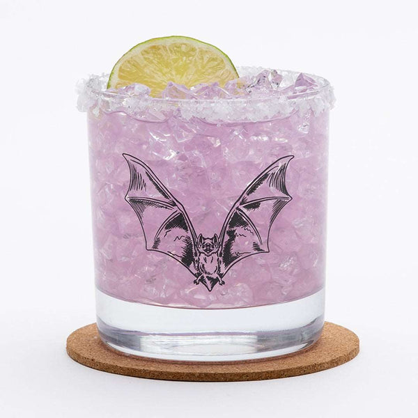 Bat illustration screen-printed onto a whiskey glass filled with purple drink and lime garnish.