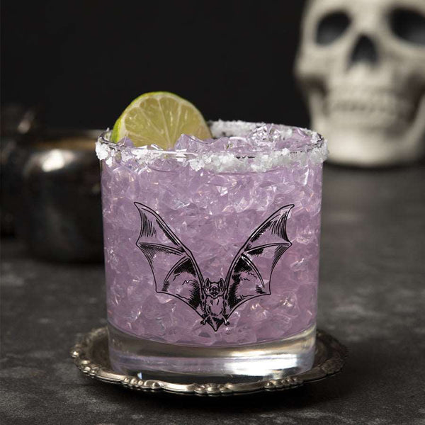 Rocks glass printed with a hand-drawn bat, printed in black ink. The glass is filled with a purple drink, ice, and a lime garnish.