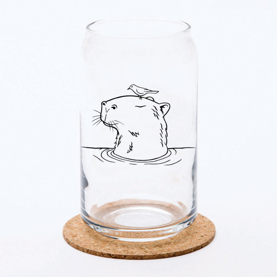 A transparent Capybara Beer Can Glass featuring an illustration of a capybara with a bird on its head, placed on a cork coaster against a simple background.