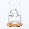 A transparent Capybara Beer Can Glass featuring an illustration of a capybara with a bird on its head, placed on a cork coaster against a simple background.