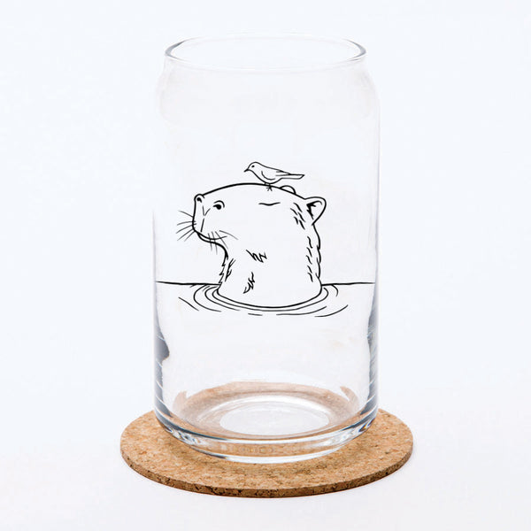 A transparent Capybara Beer Can Glass featuring an illustration of a capybara with a bird on its head, placed on a cork coaster against a simple background.