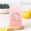 Close-up of the Capybara Beer Can Glass with a pink beverage, featuring a hand-drawn capybara illustration and a bird on its head, set on a wooden coaster with lemon slices and blueberries in the background.