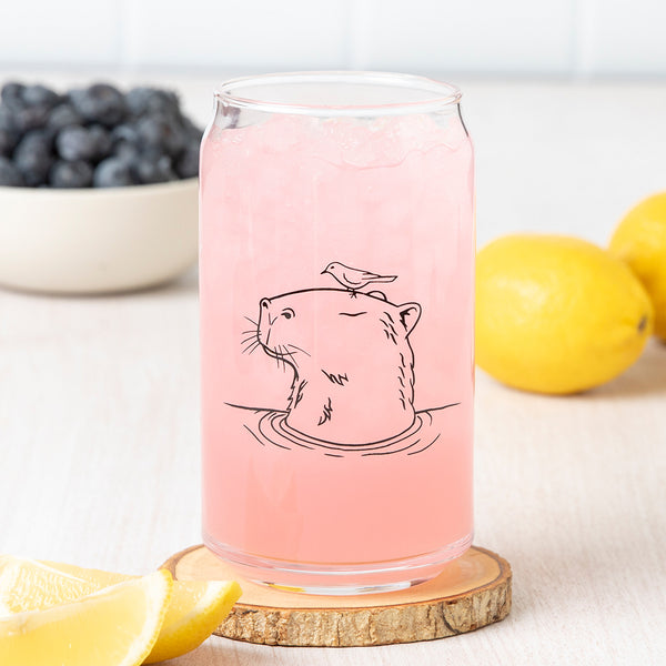 Close-up of the Capybara Beer Can Glass with a pink beverage, featuring a hand-drawn capybara illustration and a bird on its head, set on a wooden coaster with lemon slices and blueberries in the background.