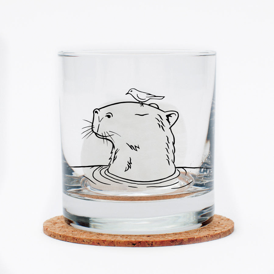 Capybara Rocks Glass featuring a black outline of a capybara with a bird on its head, displayed on a cork coaster.