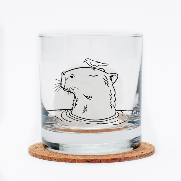Capybara Rocks Glass featuring a black outline of a capybara with a bird on its head, displayed on a cork coaster.