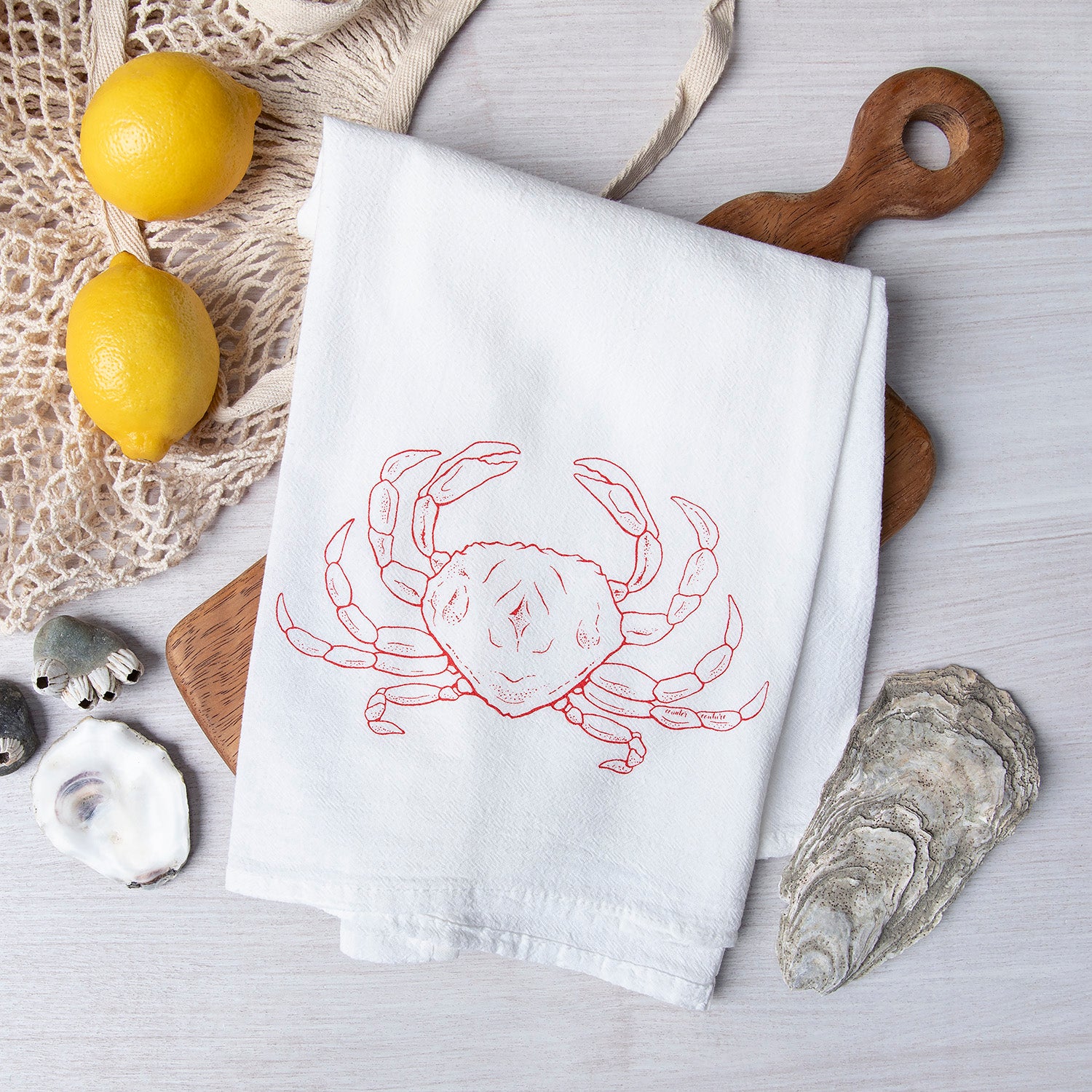 Dungeness Crab flour sack towel by Counter Couture, featuring a hand-drawn red crab illustration, styled in a kitchen setting with lemons and ocean-themed decor.