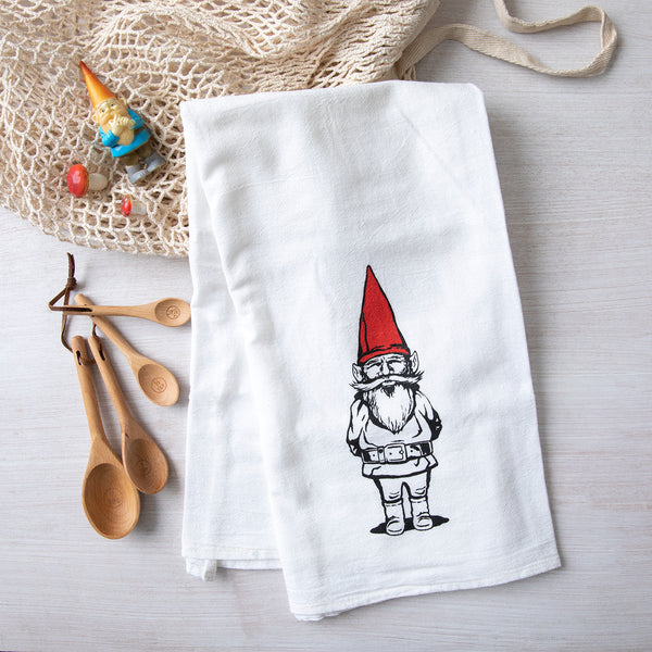 Garden gnome flour sack towel by Counter Couture, showing a whimsical gnome illustration with a red hat and beard on a white towel, placed next to wooden kitchen utensils.
