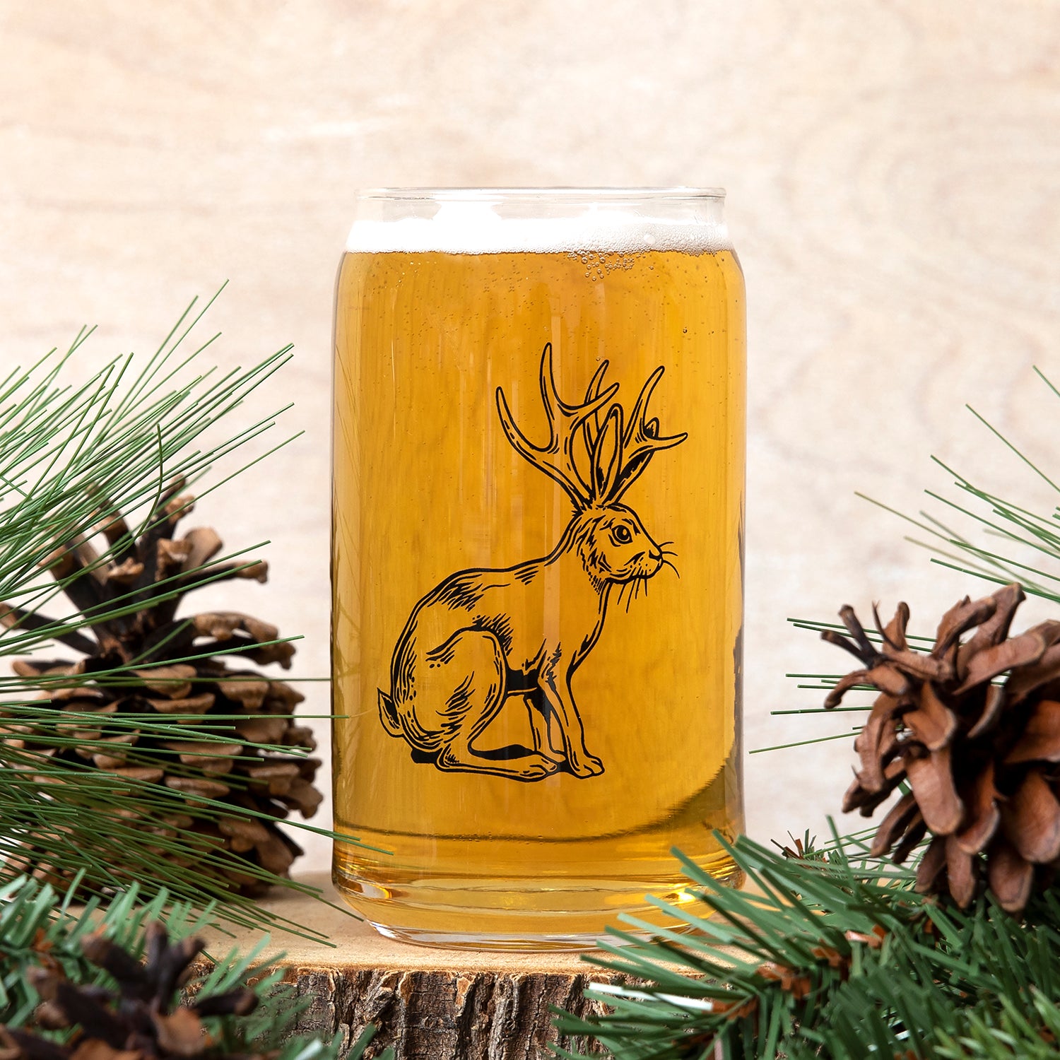 Visually similar image of Jackalope Beer Can Glass from Counter Couture, featuring a detailed jackalope illustration with beer inside, framed by pine foliage and cones.