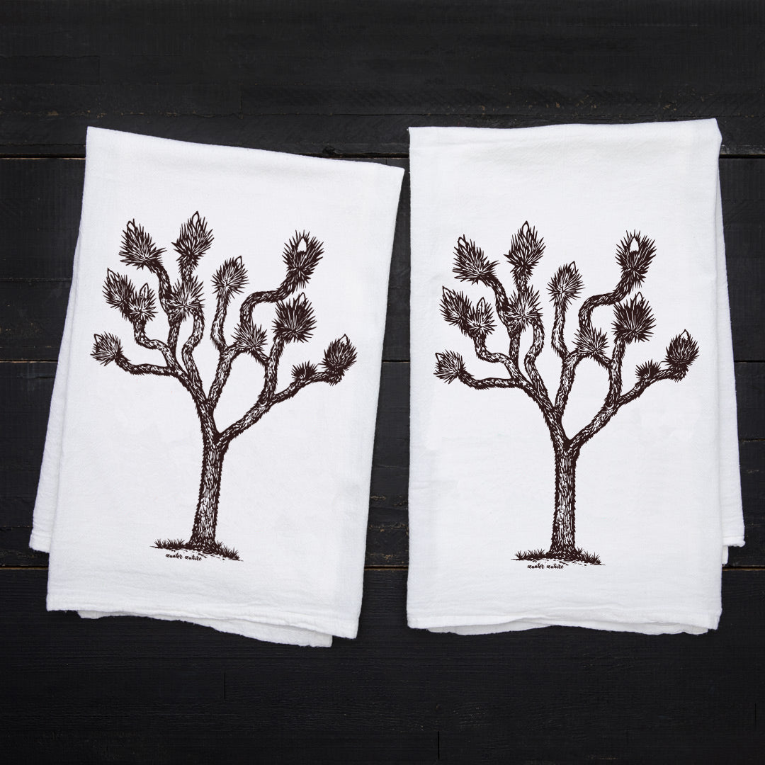 Two Counter Couture Joshua Tree flour sack towels laid flat on a dark wood surface, featuring the same hand-drawn tree design.