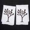 Two Counter Couture Joshua Tree flour sack towels laid flat on a dark wood surface, featuring the same hand-drawn tree design.