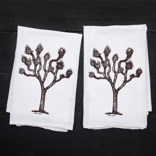 Two Counter Couture Joshua Tree flour sack towels laid flat on a dark wood surface, featuring the same hand-drawn tree design.