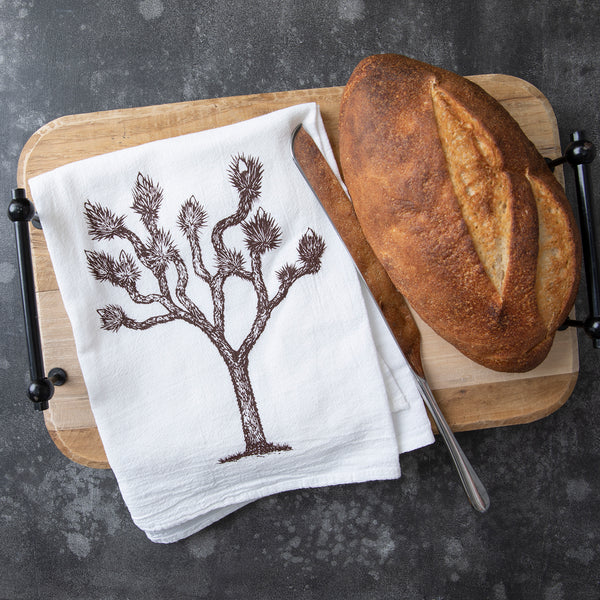 Counter Couture Joshua Tree flour sack towel displayed on a wooden cutting board with freshly baked bread.