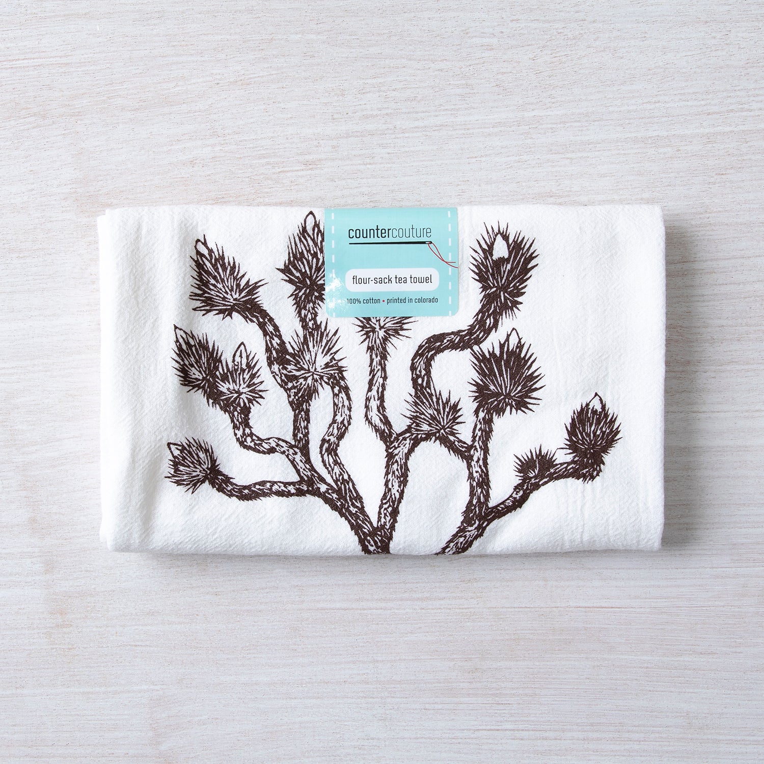 Counter Couture Joshua Tree flour sack towel wrapped with a label, placed on a light-colored wooden table.
