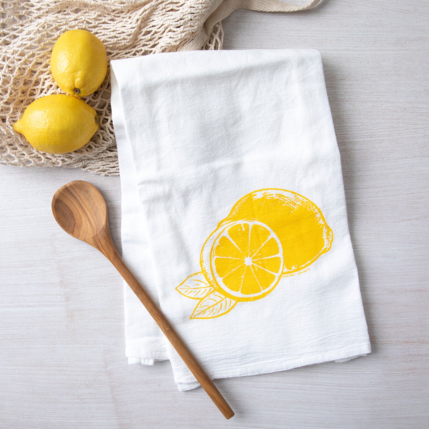 Bright yellow lemon illustration featured on a white cotton flour sack towel by Counter Couture, displayed with wooden spoon and fresh lemons in the background.