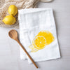 Bright yellow lemon illustration featured on a white cotton flour sack towel by Counter Couture, displayed with wooden spoon and fresh lemons in the background.