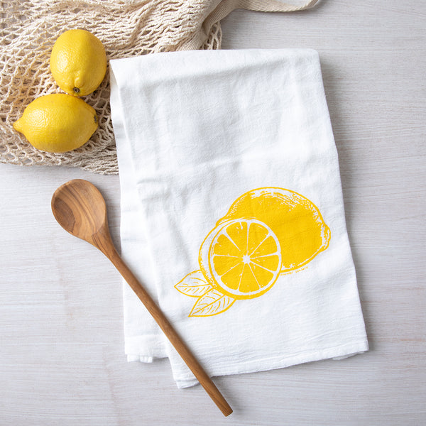 Bright yellow lemon illustration featured on a white cotton flour sack towel by Counter Couture, displayed with wooden spoon and fresh lemons in the background.