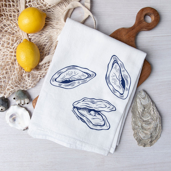 Oyster-themed cotton flour sack towel from Counter Couture, featuring hand-drawn illustrations of oysters, surrounded by lemons and decorative seashells.