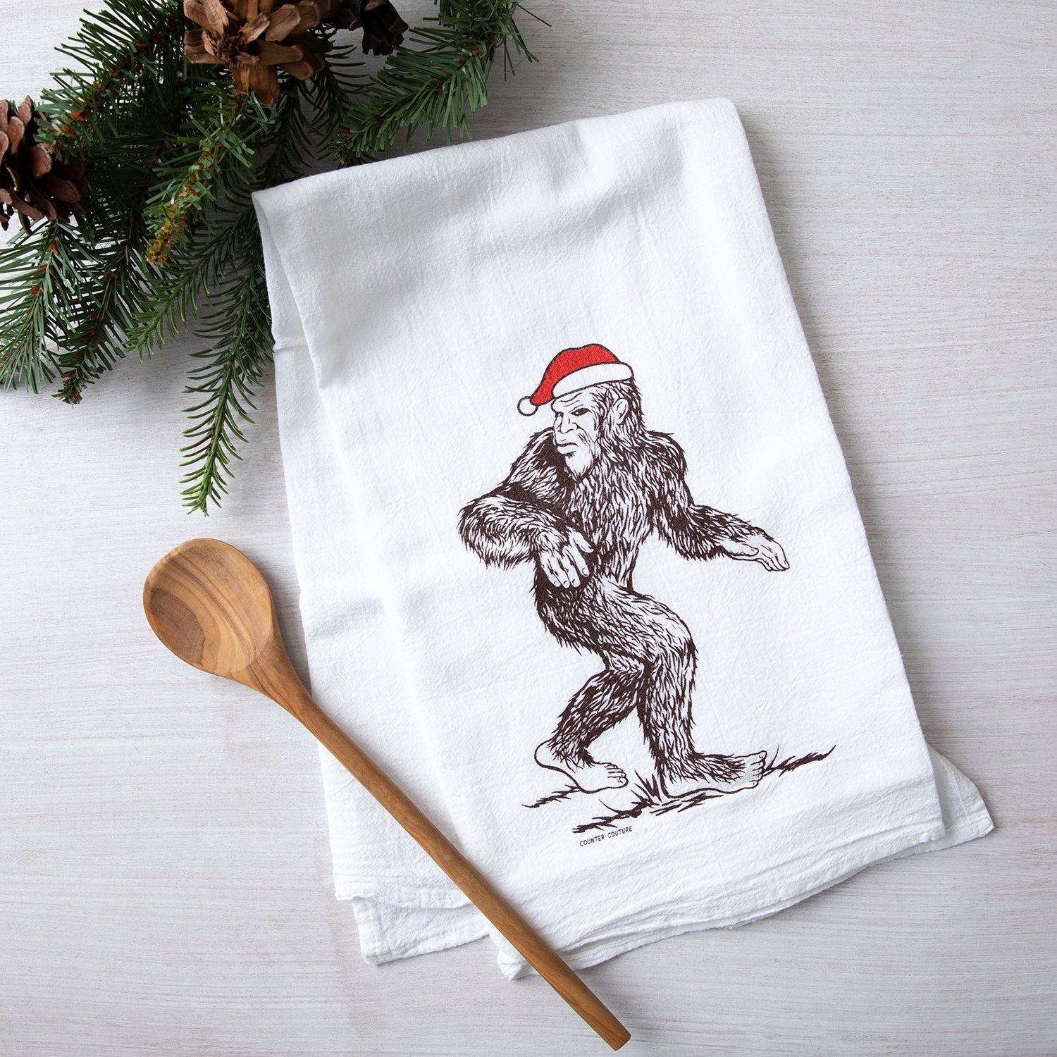 A festive Santa Sasquatch flour sack towel from Counter Couture, featuring a whimsical illustration of Sasquatch wearing a red Santa hat, placed next to a wooden spoon and pine branches.