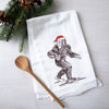 A festive Santa Sasquatch flour sack towel from Counter Couture, featuring a whimsical illustration of Sasquatch wearing a red Santa hat, placed next to a wooden spoon and pine branches.
