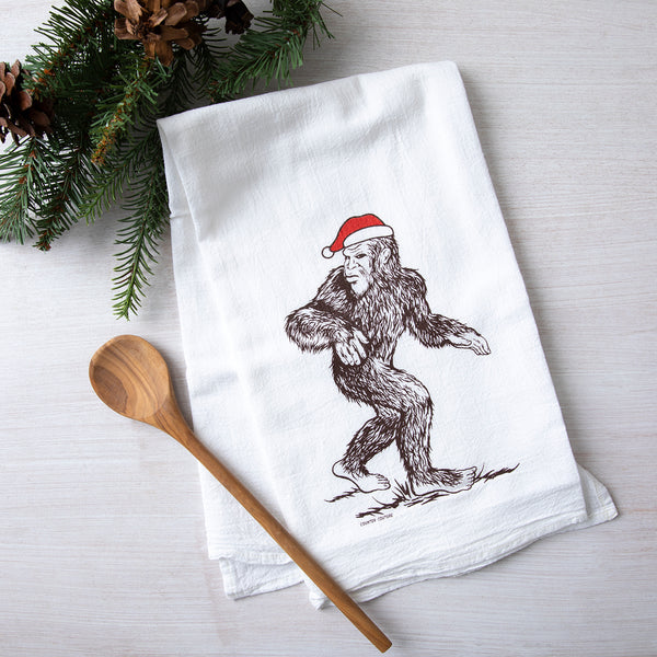 A festive Santa Sasquatch flour sack towel from Counter Couture, featuring a whimsical illustration of Sasquatch wearing a red Santa hat, placed next to a wooden spoon and pine branches.