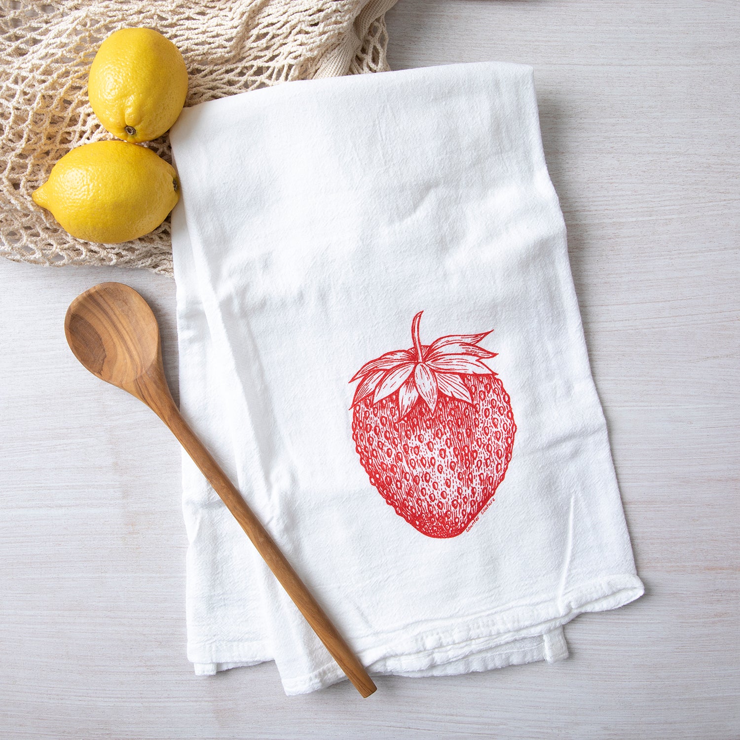 Stylish strawberry flour sack towel laid out with wooden spoon and fresh lemons, perfect for kitchen décor and functionality.