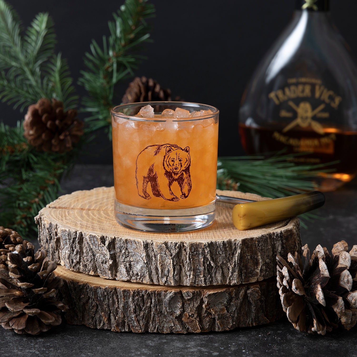 Hand-drawn bear illustration printed in brown on a whiskey glass, surrounded by pine boughs.