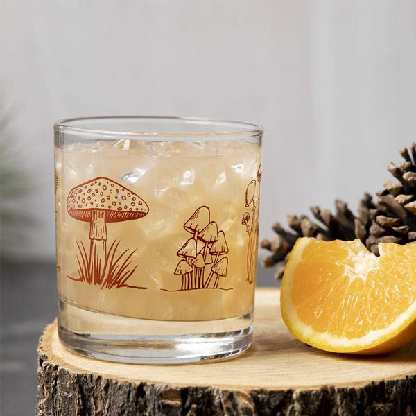 Whiskey glass with mushroom illustration, iced drink, orange slice and pinecone on wood