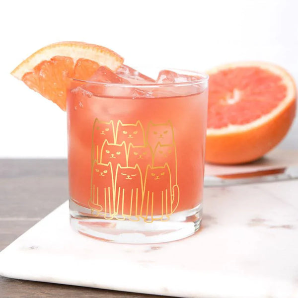 Whiskey glass with gold cat design filled with pink cocktail, ice, and grapefruit slice