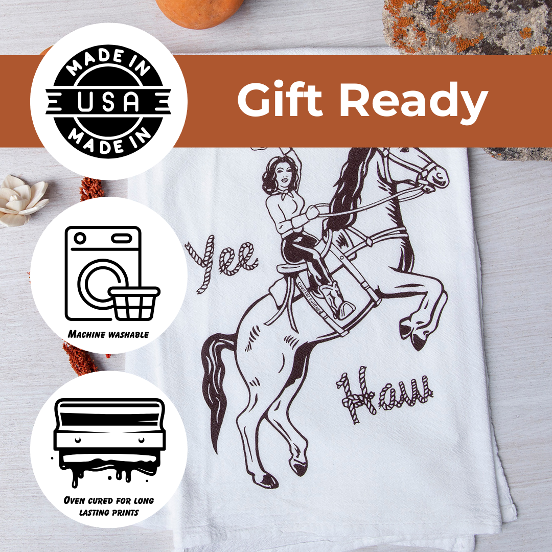 The image is a gift ready dish towel with a cartoon illustration of a woman riding a horse. The towel is displayed on a wooden surface, and there are some other items in the background.