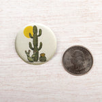 A button featuring a cactus illustration next to a United States quarter coin on a wooden surface.