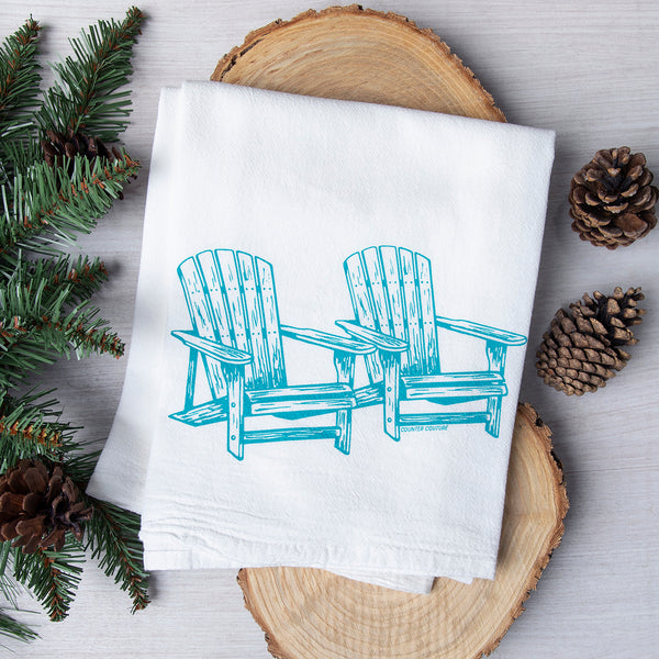 Adirondack Flour Sack Towel by Counter Couture featuring a hand-drawn illustration of two Adirondack chairs in aqua blue, set against a rustic background with pine branches and pine cones.