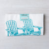 Adirondack Flour Sack Towel by Counter Couture, neatly folded with a label, showcasing a hand-drawn design of two Adirondack chairs in aqua blue on a light wood surface.
