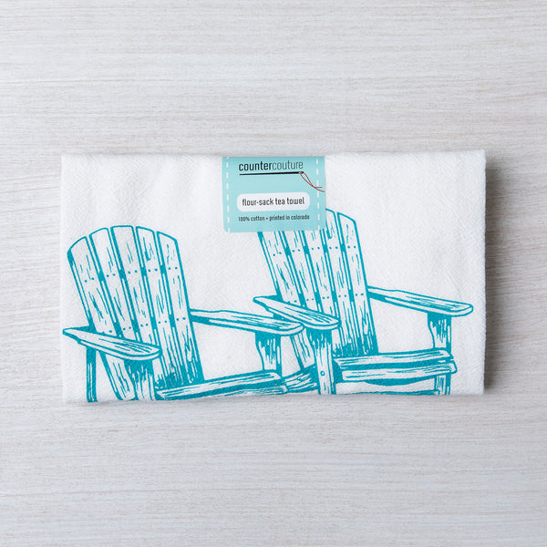 Adirondack Flour Sack Towel by Counter Couture, neatly folded with a label, showcasing a hand-drawn design of two Adirondack chairs in aqua blue on a light wood surface.