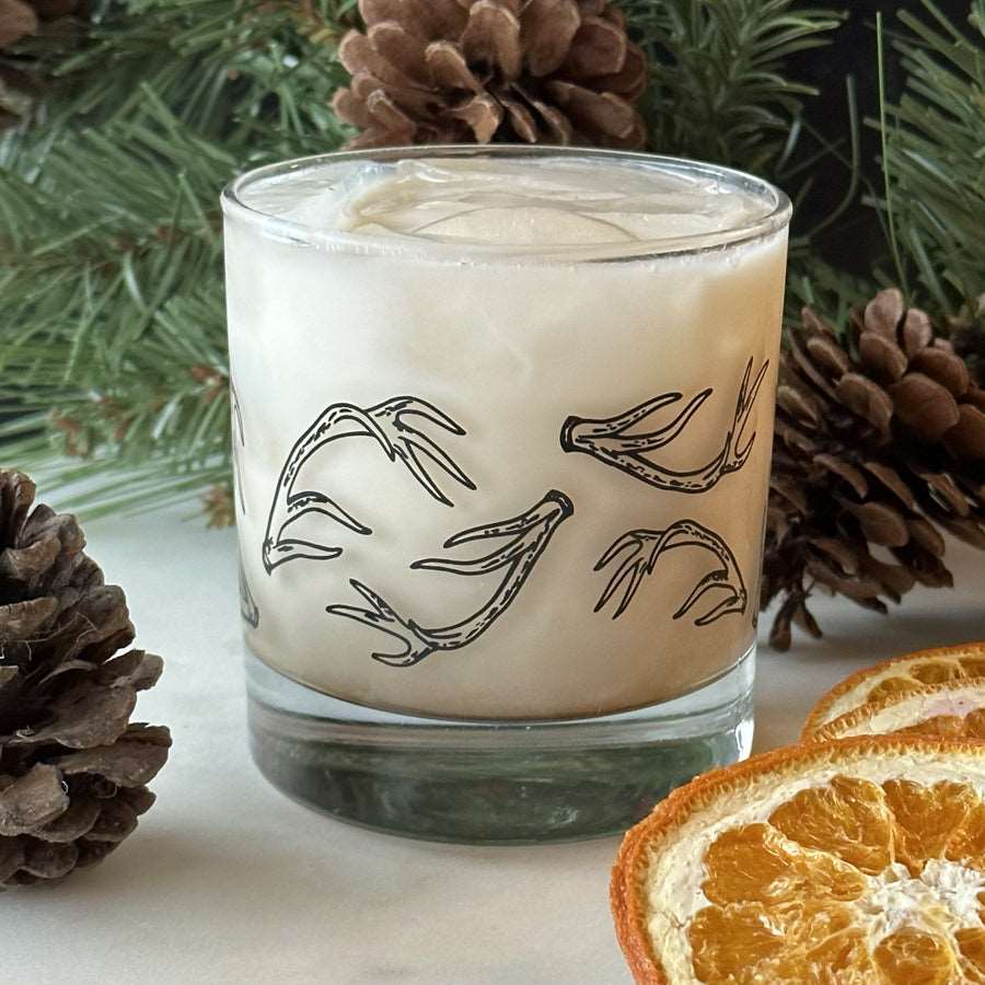 A rocks glass filled with a white drink and ice is printed with black hand-drawn antlers. Pine boughs and dried oranges for decoration.