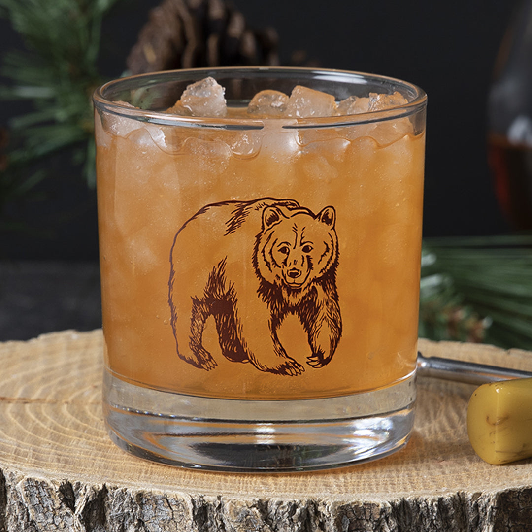 Close-up of a bourbon cocktail in a glass with a bear illustration, on a wood slice, rustic barware