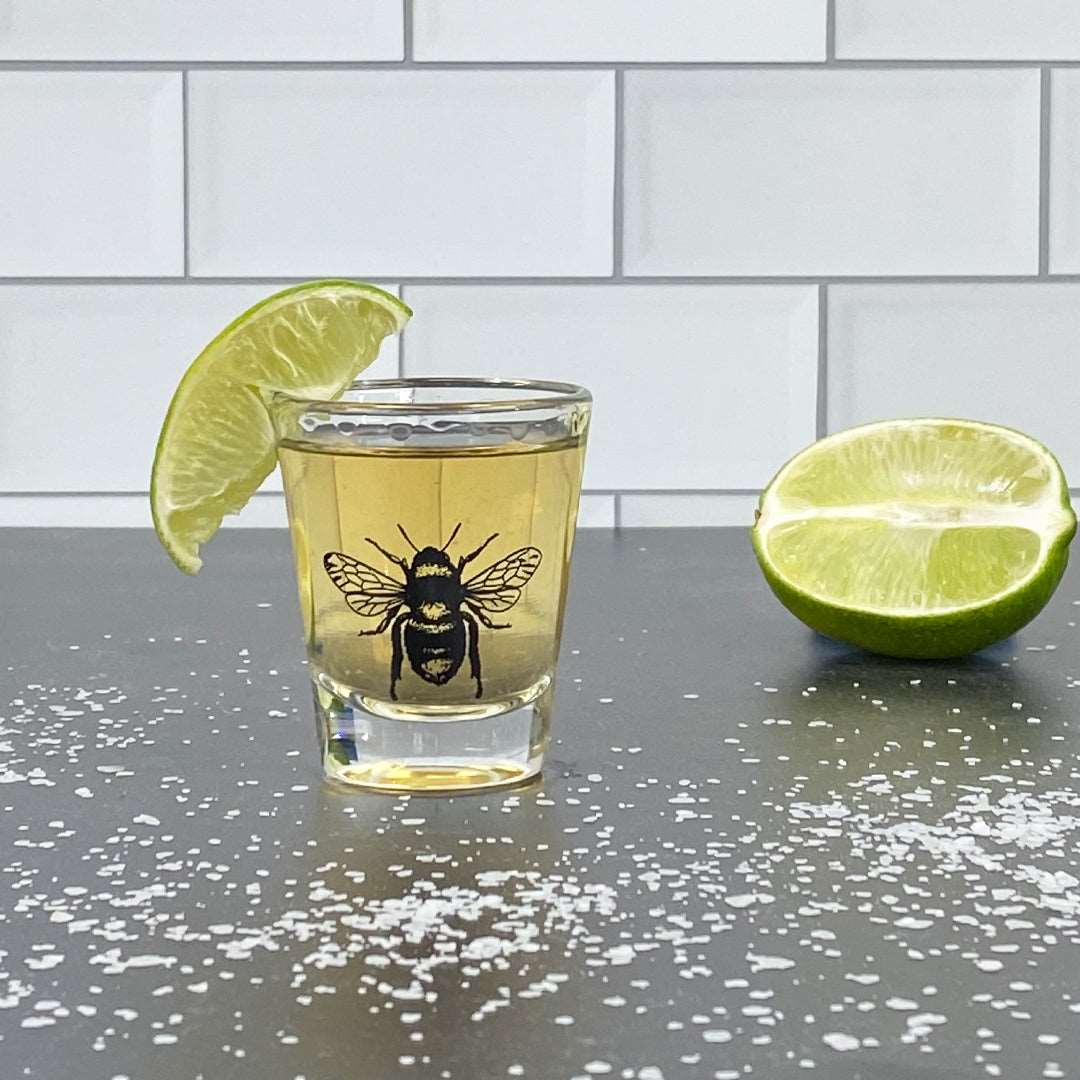 Bee shot glass with tequila, lime wedge, and salt on black counter with tile backdrop