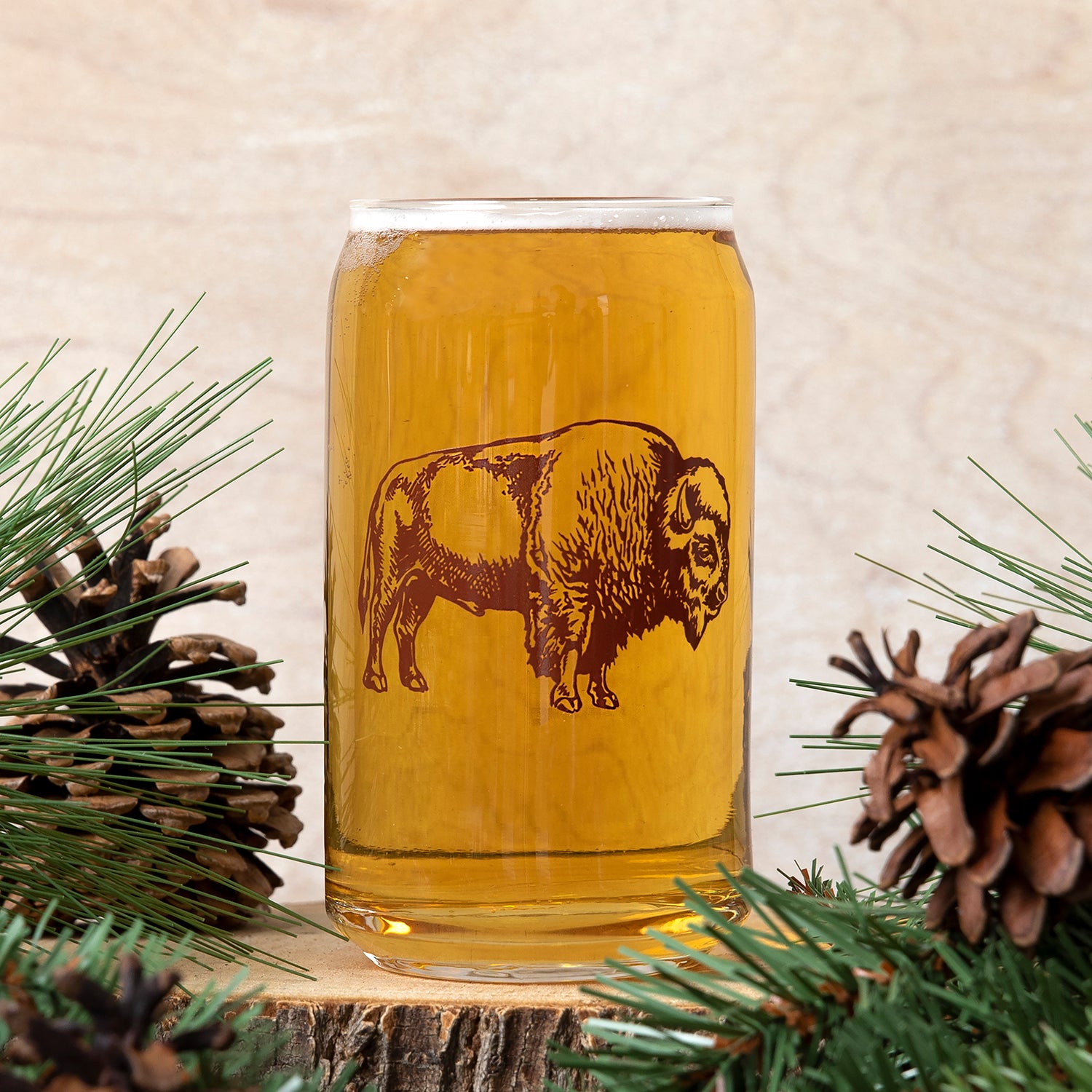 Bison print can-shaped beer glass with amber drink, pine branches and pinecones accent