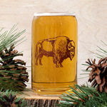 Closeup of a beer can glass with a brown bison graphic, surrounded by pinecones and greenery.