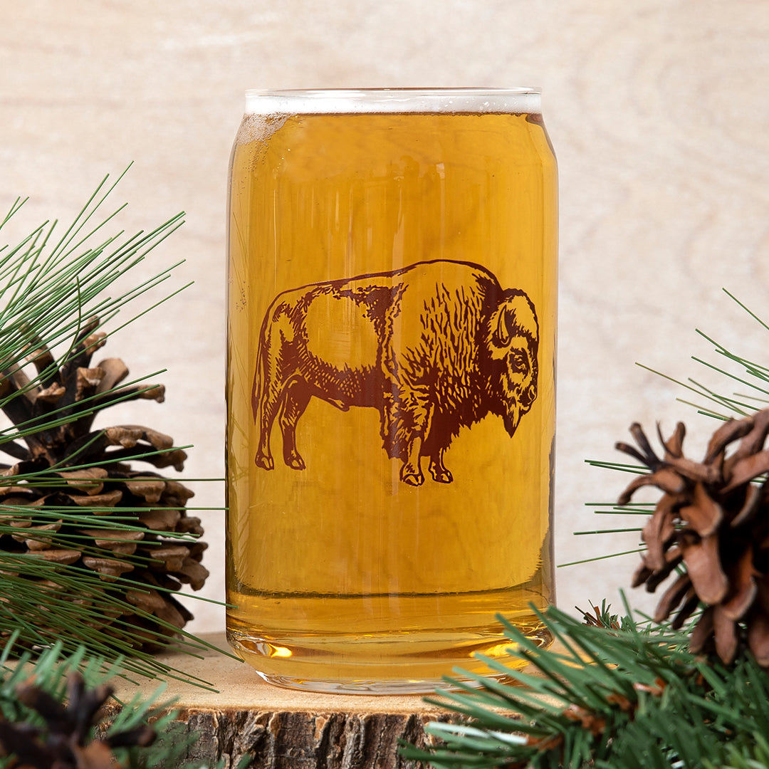 Closeup of a beer can glass with a brown bison graphic, surrounded by pinecones and greenery.