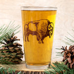 Pint glass with bison graphic filled with beer, surrounded by pinecones and pine branches.