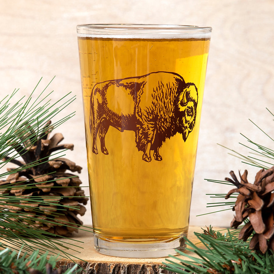 Pint glass with bison graphic filled with beer, surrounded by pinecones and pine branches.