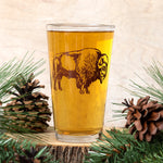 Pint glass with brown bison illustration, filled with beer, styled with pine and pinecones.