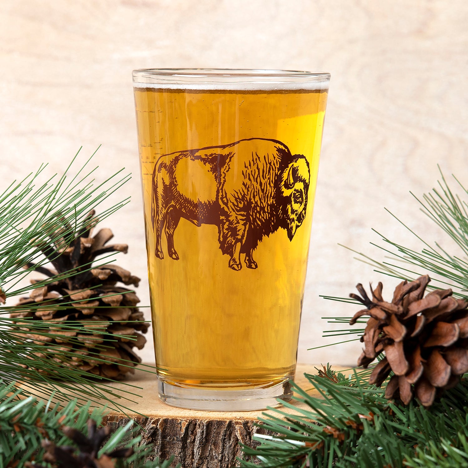 Pint glass with brown bison illustration, filled with beer, styled with pine and pinecones.