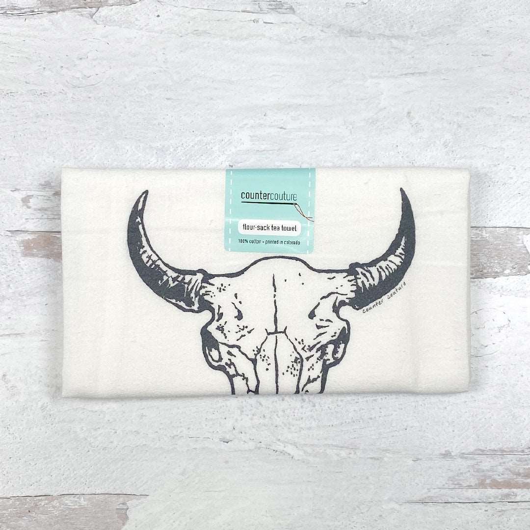 Flour sack tea towel with black buffalo skull illustration, folded and packaged on white wood.