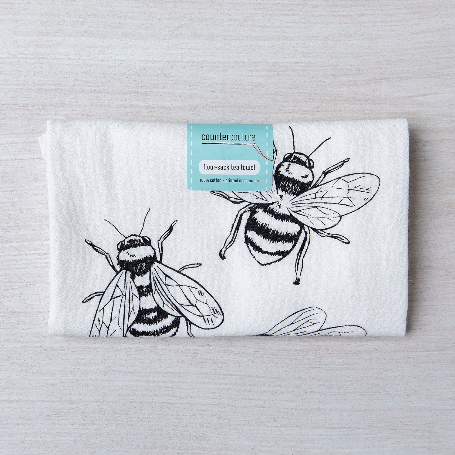 Folded Bumble Bee flour sack tea towel displaying intricate bee designs, with a tag indicating it is made of 100% cotton and printed in Colorado.