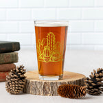 Pint glass with yellow cactus design, filled with beer, staged on wood with pinecones