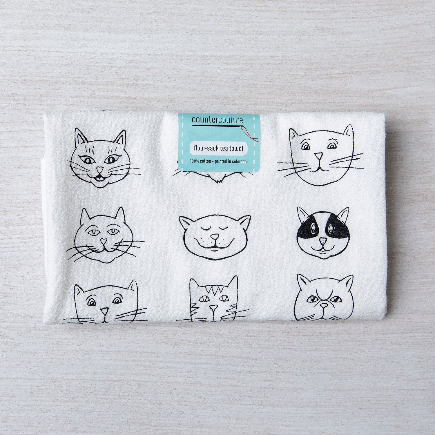 Folded Cute Cat Faces Flour Sack Towel by Counter Couture with a label displaying its eco-friendly material, featuring playful hand-drawn cat illustrations, ideal for kitchen decor.
